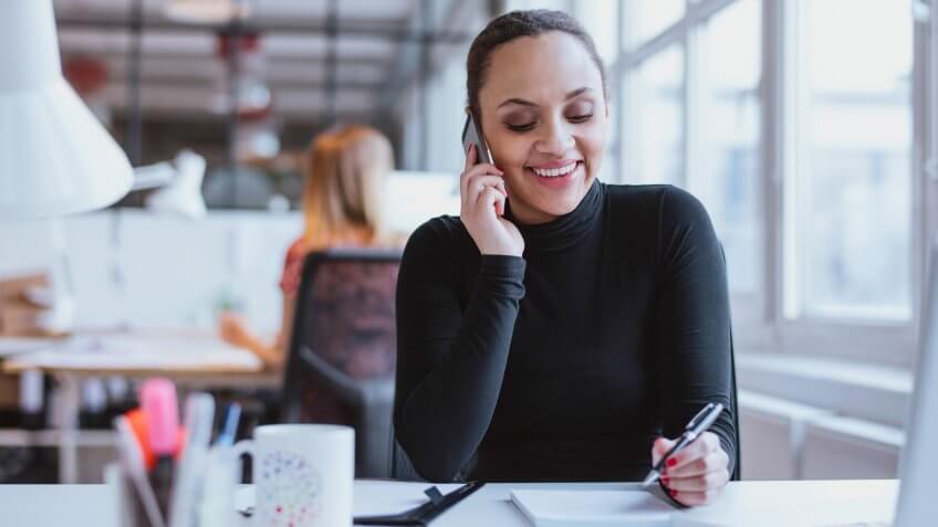 Happy young woman sitting at her desk working and answering a phone call.