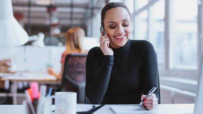 Happy young woman sitting at her desk working and answering a phone call.