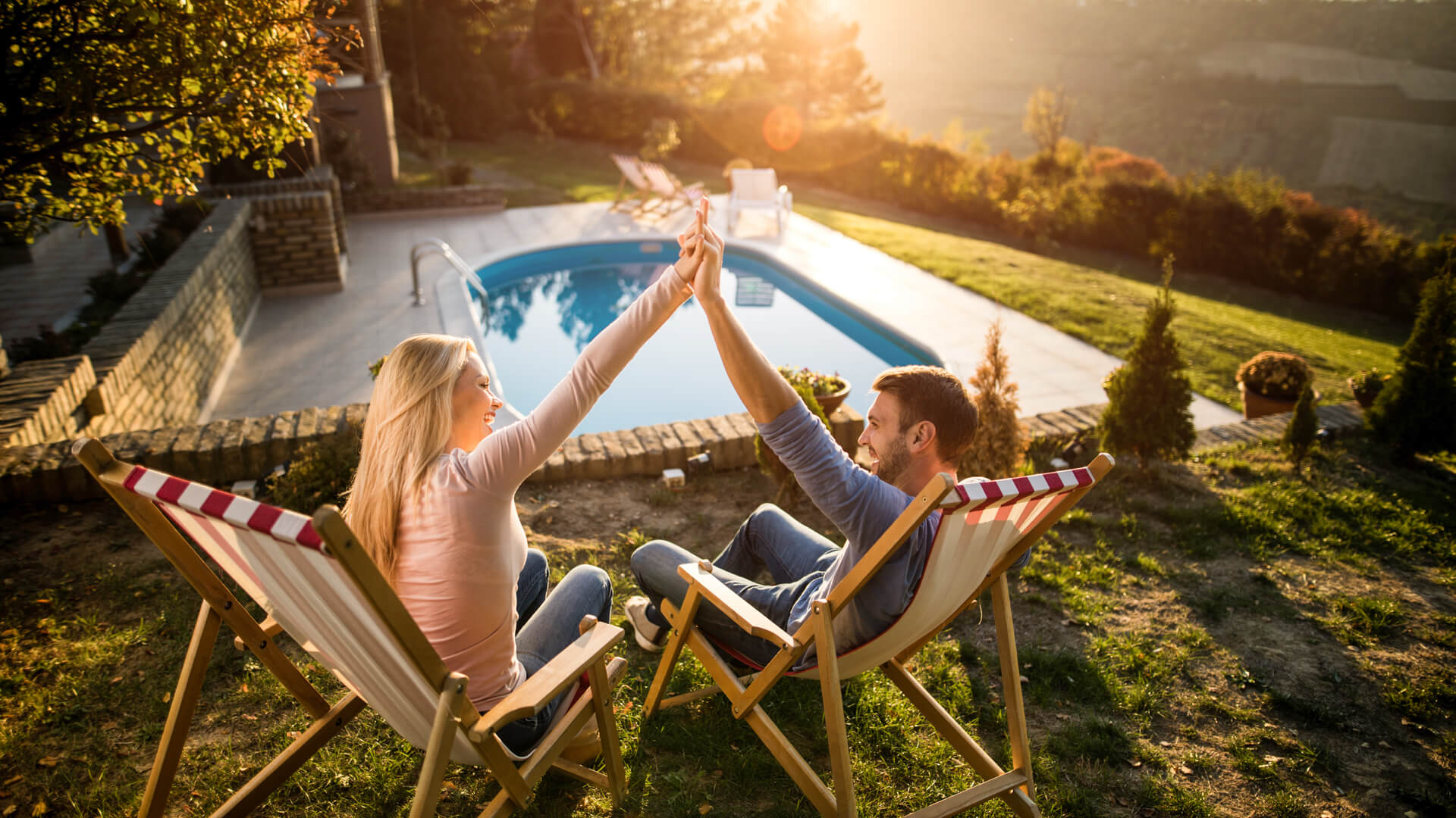 Young happy couple relaxing in deck chairs at their back yard and holding their arms together.