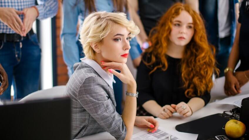 close up portrait of serious elegant secretary with fair hair holding her hand on chin at workplace among co-workers.