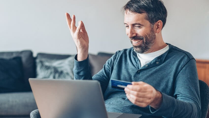 Smiling man holding a credit card and using laptop at home.