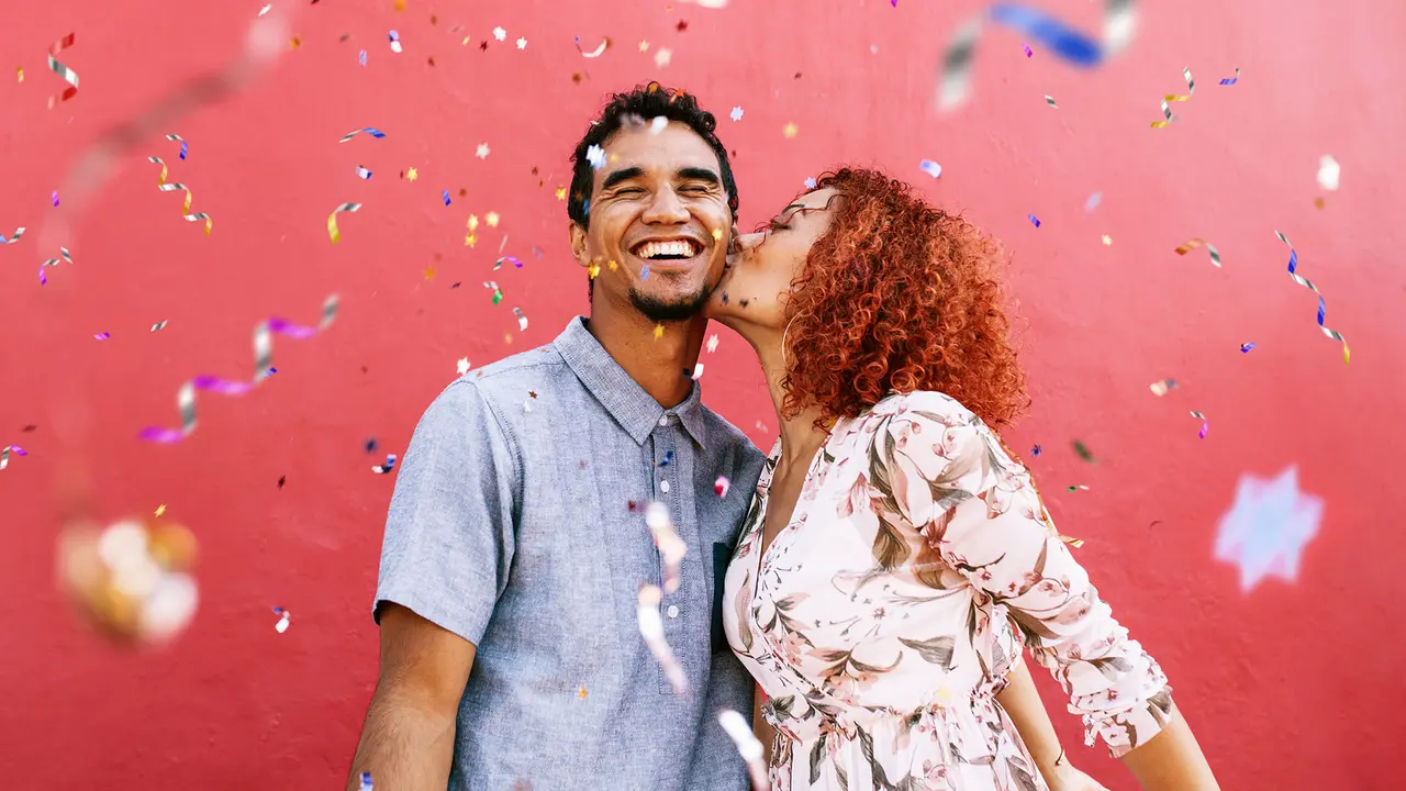 Young woman kissing man on cheek with confetti falling all around them.