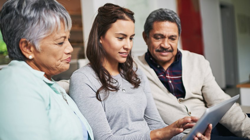 Shot of a young woman showing something to her parents on her tablet while they sit on a sofa together.
