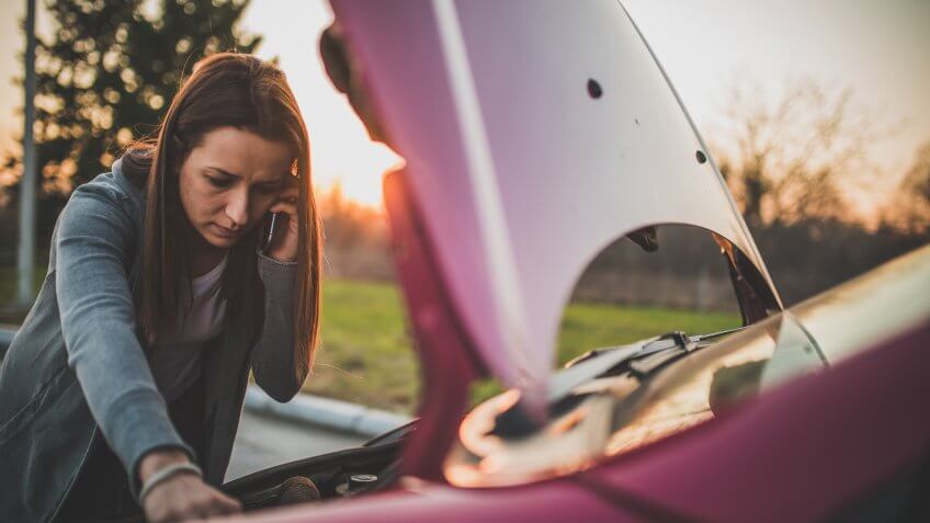 Young woman standing by her broken car on the road and using phone to call for help.