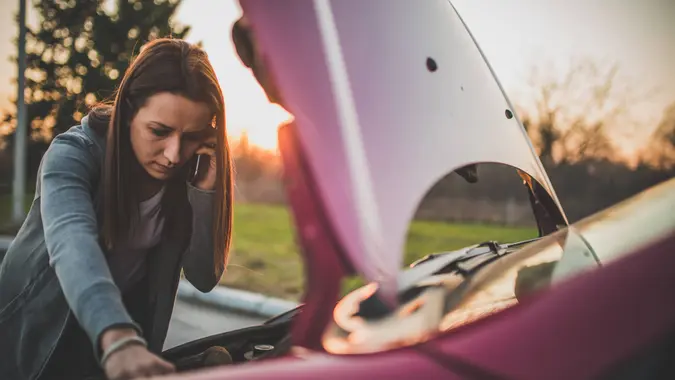 Young woman standing by her broken car on the road and using phone to call for help.