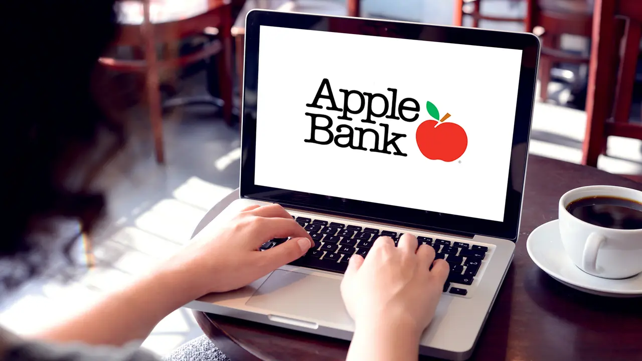 Woman sitting down in a cafe with a cup of coffee and using a laptop with the Apple Bank website pulled up on it.