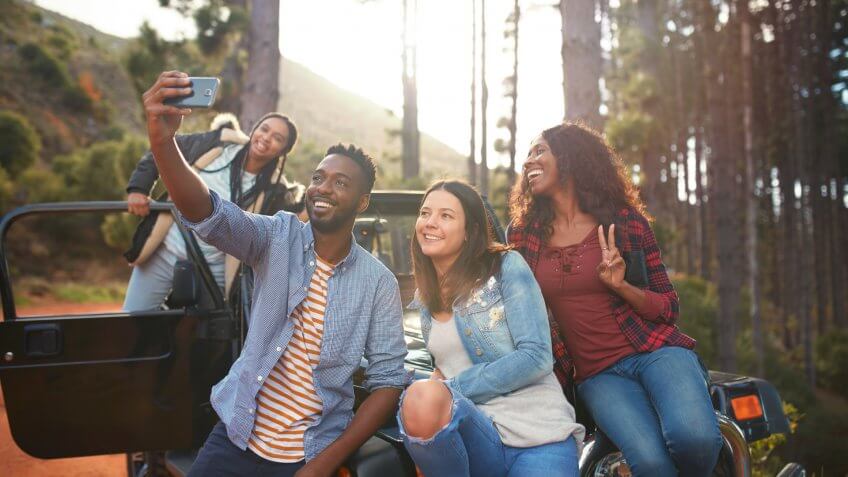 Young friends with camera phone taking selfie at jeep in woods.