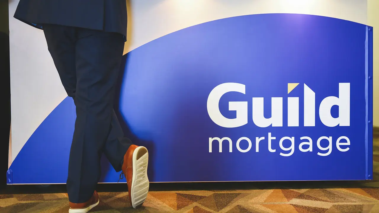 Man in navy suit and blue shoes stands facing an indoor Guild Mortgage board, back toward the lens