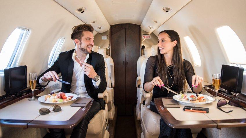Young well dressed man sitting inside private aeroplane, holding glass of champagne and looking outside the window.