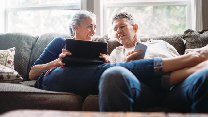 A couple in their 50's relax in their home on the living room couch, enjoying reading and surfing the internet on their mobile touchscreen phones and computer tablet.