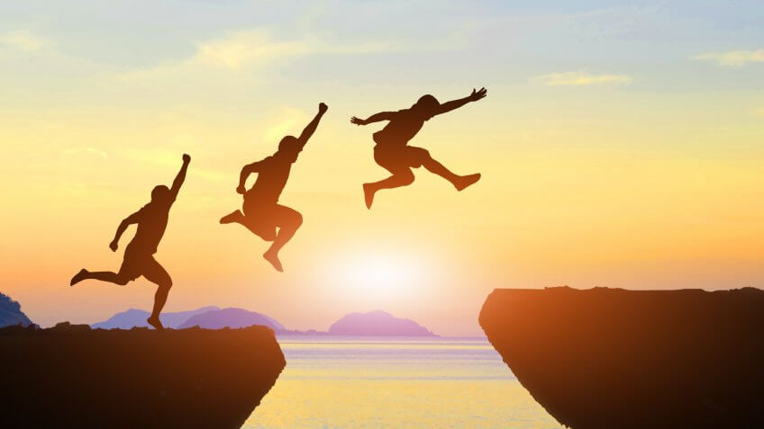 Men jump cliff sun light over silhouette.