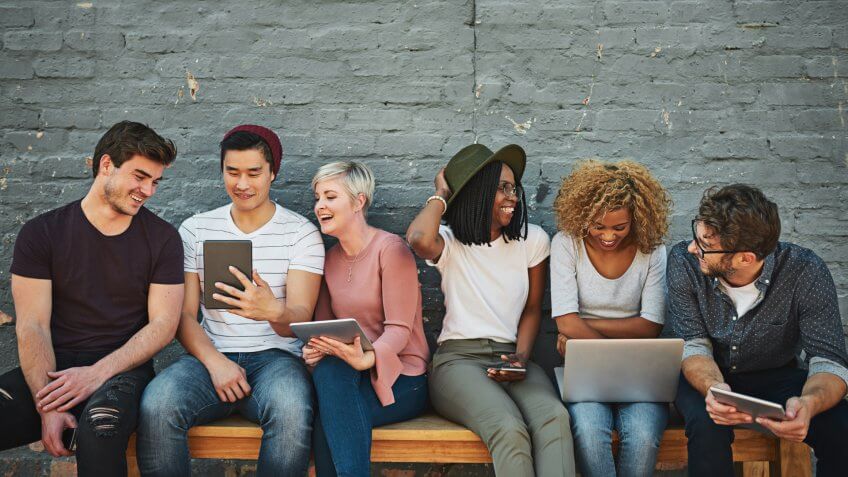 Millennial friends sitting on a bench viewing tablets and laptop