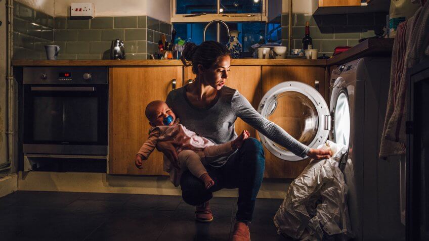 Young mother is holding her crying baby daughter on her hip while trying to load the washing machine.
