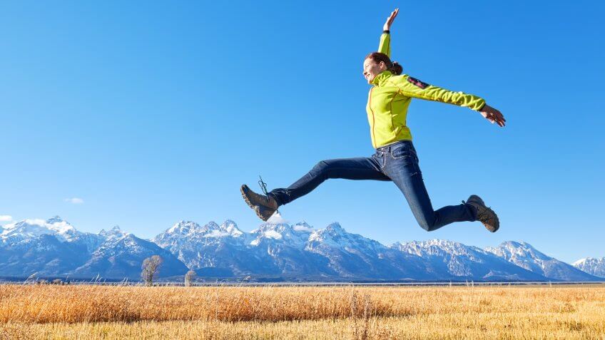 Happy young woman jumping on a meadow, Grand Teton mountain range in distance, Wyoming, USA.
