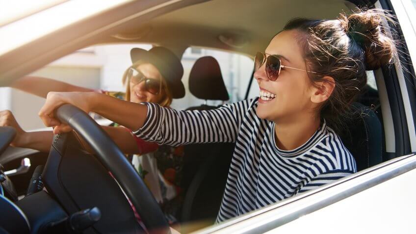 Two attractive young girlfriends wearing sunglasses talking and laughing together while driving in a car through the city on a sunny day.