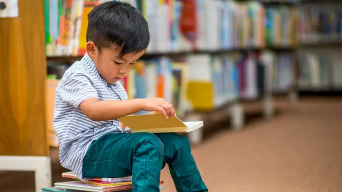 A little boy is sitting on a stack of books at the library and is reading.