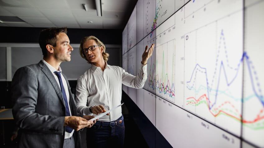 Group of business professionals in a dark room standing in front of a large data display screen with information.