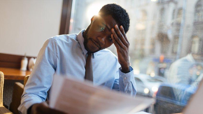 Focused African-American office manager sitting at cafe table with laptop, reading important documents with puzzled expression and holding head with his hand.