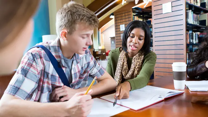 Teen or young adult African American college girl is tutoring a teenage Caucasian high school boy while they sit at table in modern school or public library.