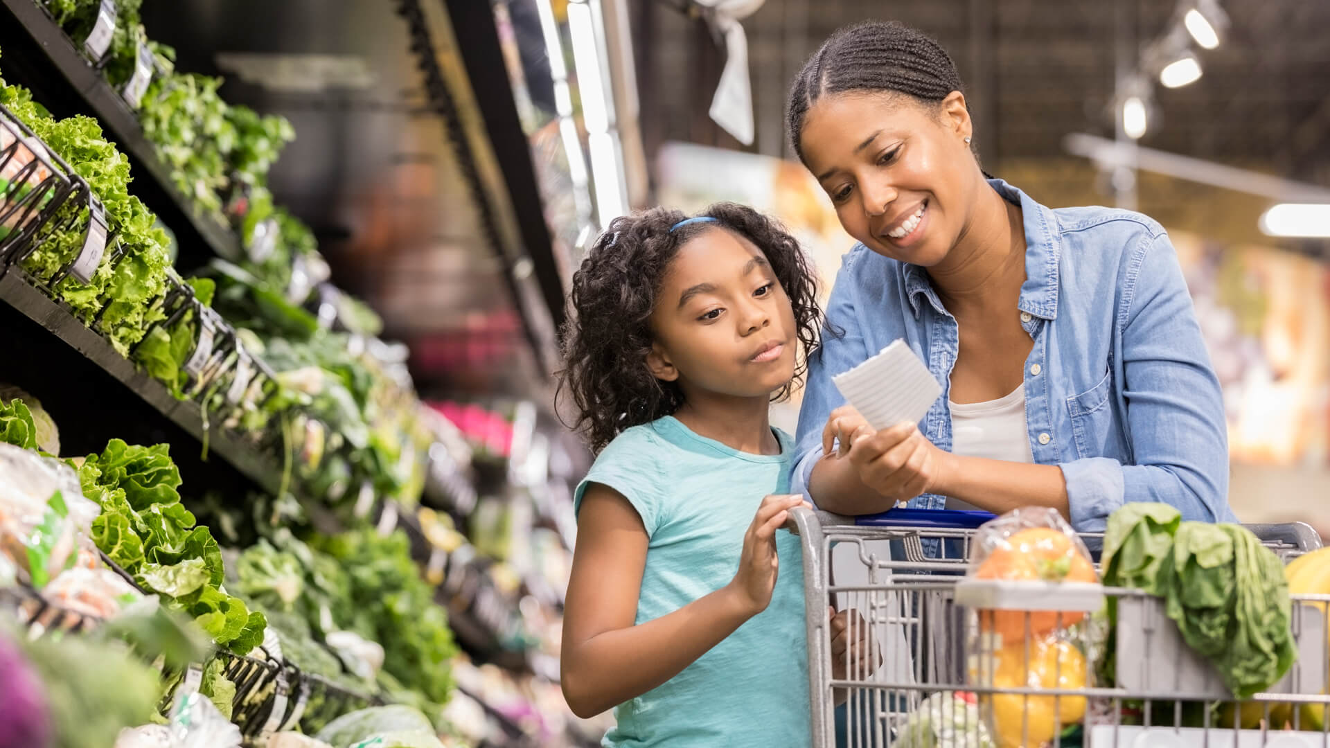 A smiling mid adult woman stands in the produce section of her supermarket and reaches down to show her serious elementary age daughter the next items on a paper shopping list.