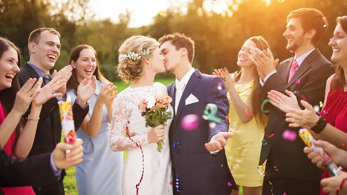 Full length portrait of newlywed couple and their friends at the wedding party showered with confetti in green sunny park.