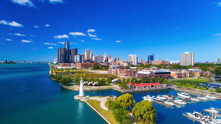 Aerial with Downtown Detroit skyline in the distance, with a blue sky with puffy fair weather clouds and Windsor, Ontario, Canada in the background, the Detroit River on the left, and a lighthouse, marina, and park in the foreground.