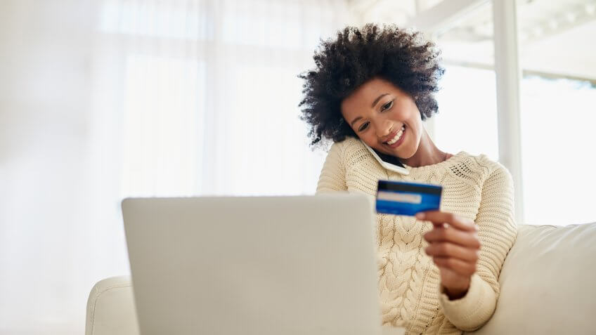young woman on her phone holding credit card while using her laptop while sitting on the sofa at home