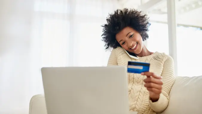 young woman on her phone holding credit card while using her laptop while sitting on the sofa at home