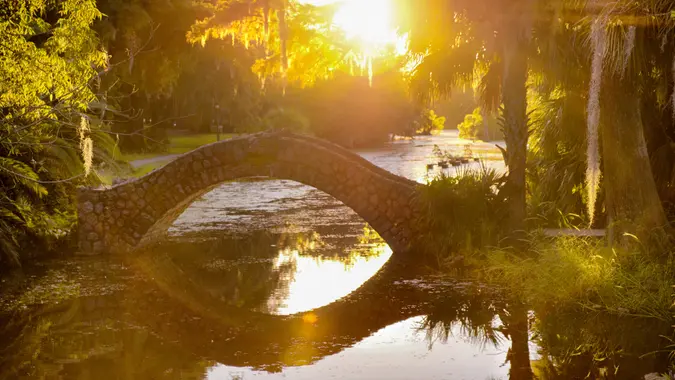 Rustic stone bridge arching over the still water in City Park in New Orleans, Louisiana at dusk with palm trees and Cypress trees.