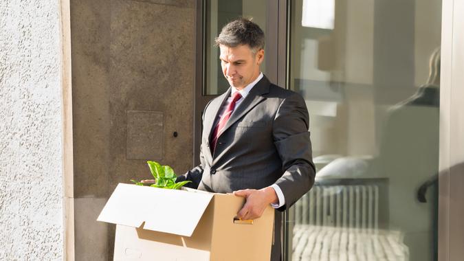 Sad Mature Businessman Moving Out With Cardboard Box From Office.