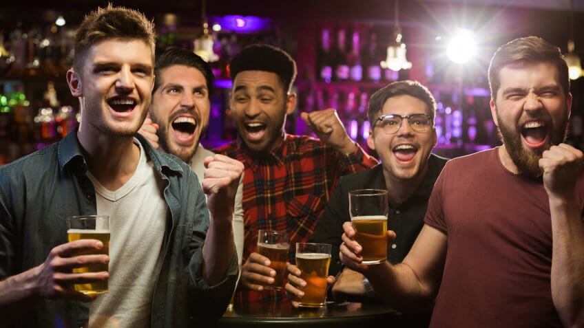 Four happy men holding beer mugs and gesturing while watching TV in bar or pub.