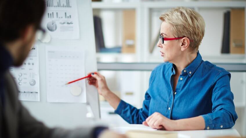 Experienced financier pointing at table with financial data on whiteboard while explaining it to young co-worker