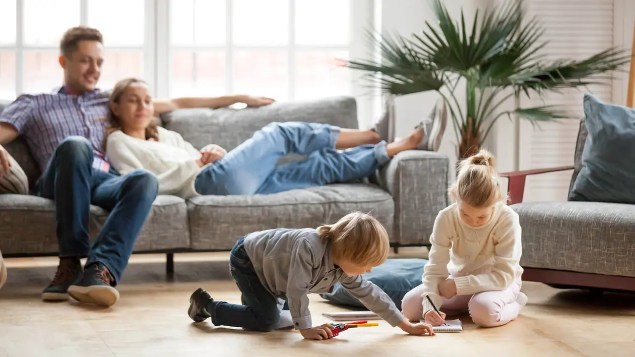 Children sister and brother playing drawing together on floor while young parents relaxing at home on sofa, little boy girl having fun, friendship between siblings, family leisure time in living room.