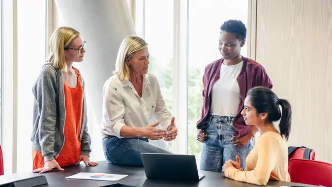 Multi racial group of female students listening to college lecturer with laptop on table.