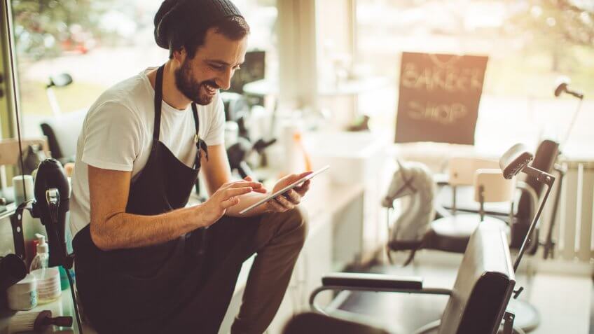 Photo of a young barber, sitting proudly on a counter in his barber's shop and using a digital tablet.