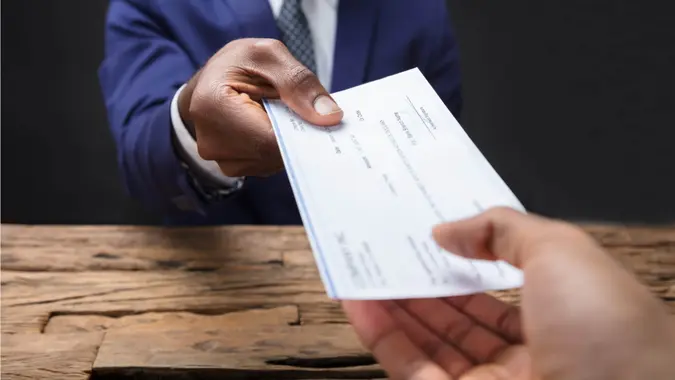 A person signs a cashier’s check with a pen while holding it over a desk with financial documents.
