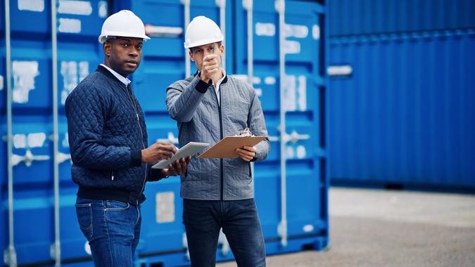 Two engineers tracking inventory using a clipboard and digital tablet while standing by freight containers on a large commercial shipping dock.