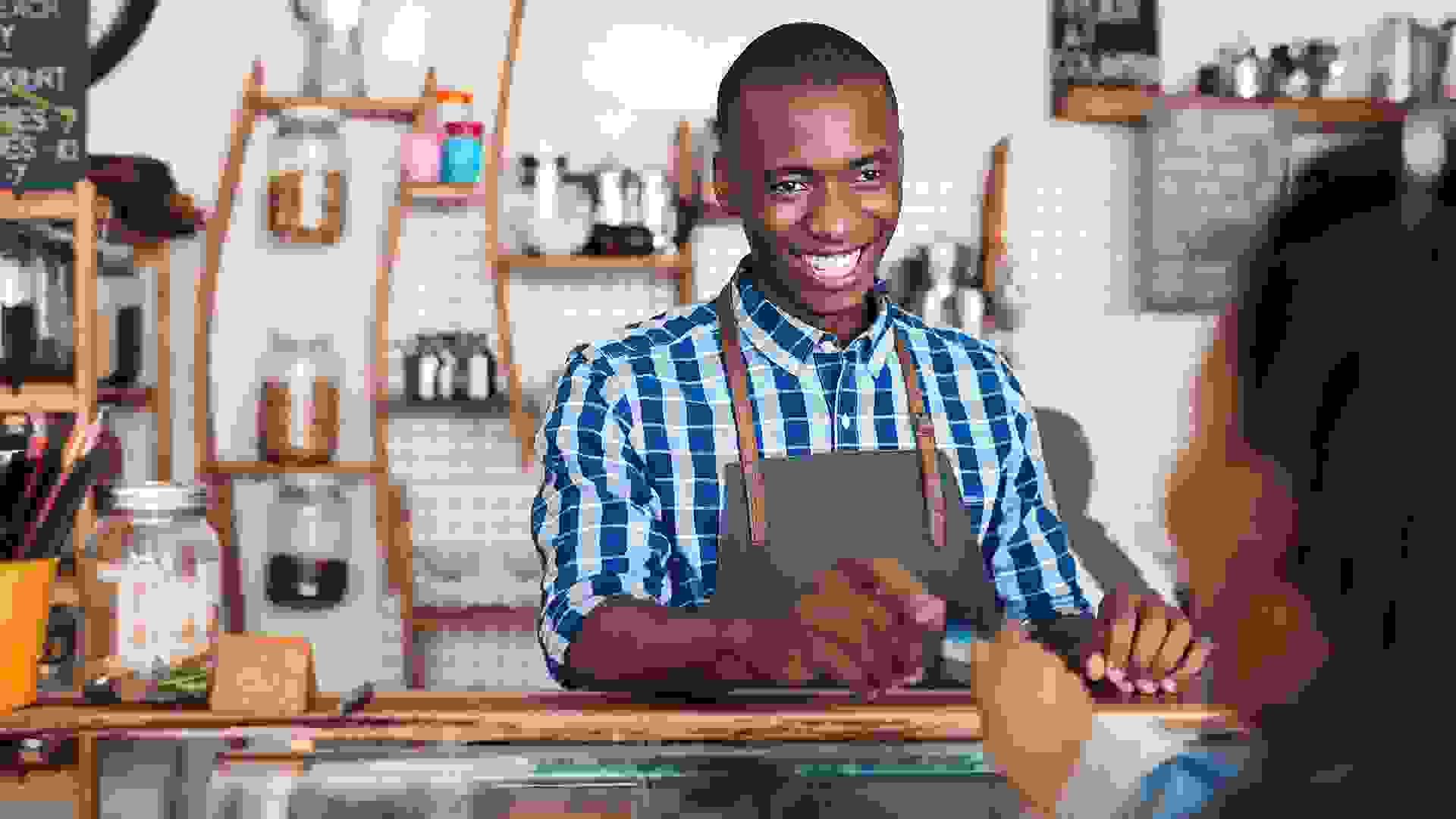 Smiling barista standing behind a counter in a cafe taking a credit card from a customer to pay for her purchase - Image.