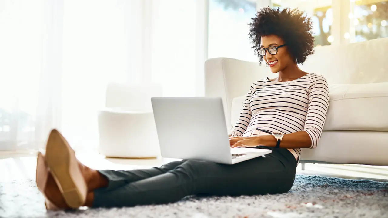 A cheerful young woman is shopping on her laptop while seated on the floor at home.