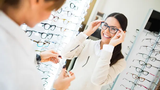 Young smiling woman is trying on eyeglasses in optical shop with the assistance of optometrist.