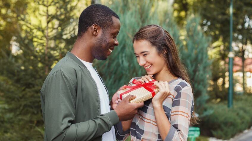 Young african-american man with giftbox surprising his girlfriend with anniversary present, walking in park.