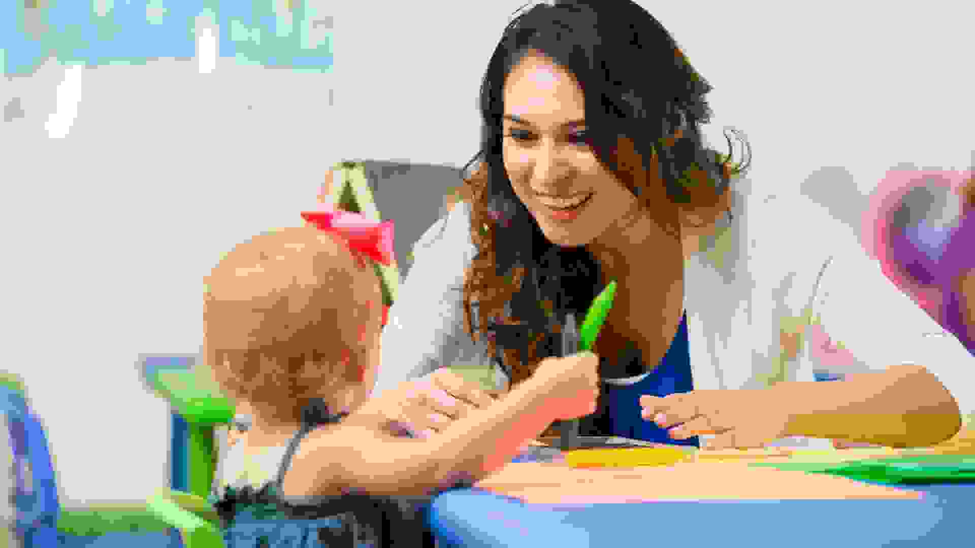 Beautiful Hispanic female daycare teacher leans on table and helps cute blonde toddler girl with school project.