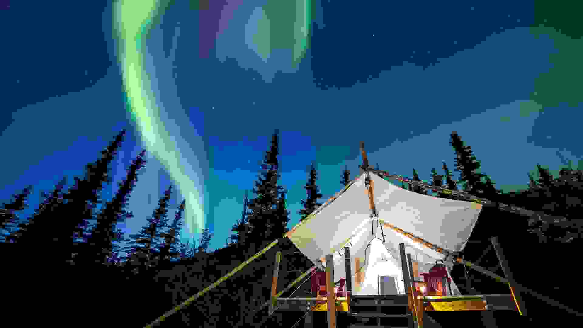 Aurora Borealis glowing green and pink over large canvas luxury camping tent in Alaska.