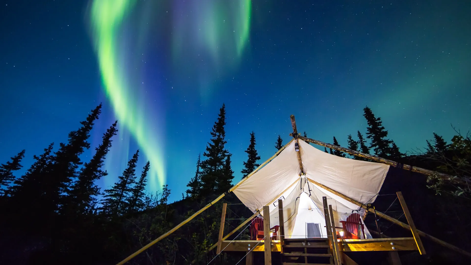 Aurora Borealis glowing green and pink over large canvas luxury camping tent in Alaska.