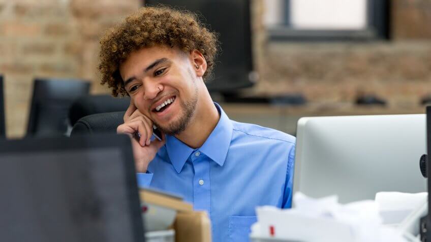Business man on the phone smiling while sat in front of his computer screen.