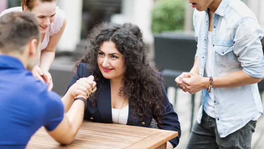 Young woman and young man arm wrestling in cafe, their friends cheering them.