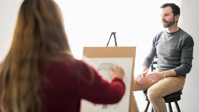 Rear view of a young female artist sketching a man sitting on stool indoors