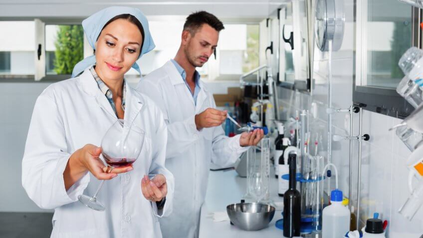 Two young efficient researchers in white coat checking wine acidity and sugar saturation in chemical laboratory.