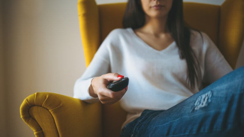 Close up of woman in armchair with tv remote control changing television channels.