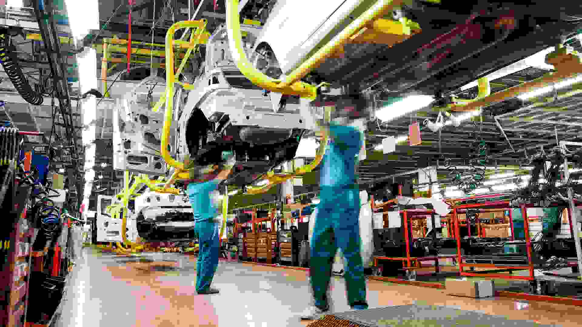 Two workers wearing green shirts and blue pants work underneath a car in an automobile factory.